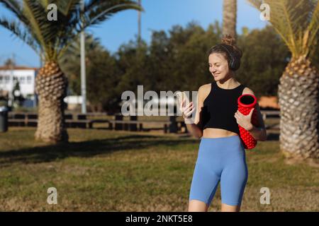 Lächelnde junge Frau macht Pause beim Training, steht mit rotem Massagerollzylinder, benutzt Smartphone, hört Lieder in Kopfhörern, trägt Sportbekleidung Stockfoto
