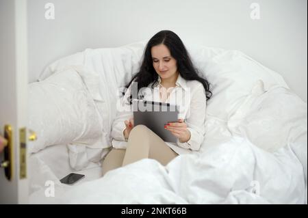 Schöne, schwarzhaarige Frau sitzt auf einem weißen Sofa neben einer Bettdecke in den Händen mit einem Tablet Frau arbeitet Spiele kommuniziert nahe gelegene Mobiltelefone Soziale Netzwerke Internet Freunde anrufen angenehme Emotionen Stockfoto