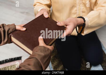 Abgeschnittene Ansicht eines muslimischen Mannes mit Gebetsperlen, der seinem Sohn zu Hause ein Buch gibt, Bild von der Börse Stockfoto