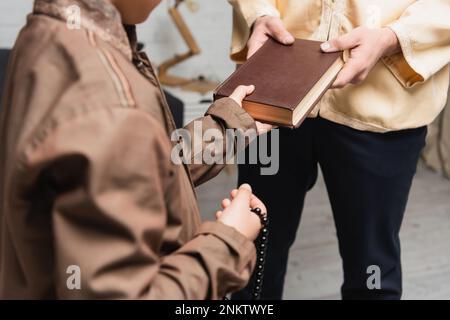 Zugeschnittener Blick auf den muslimischen Vater, der seinem Sohn ein Buch gibt, mit Gebetsperlen zu Hause, Stockbild Stockfoto