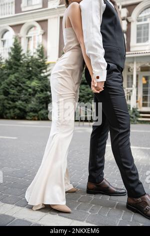 Kurzer Blick auf junge Braut und Bräutigam im Hochzeitskleid und Anzug, die sich draußen die Hände halten, Stockbild Stockfoto