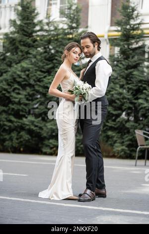 Volle Länge des bärtigen Mannes in Weste umarmt Braut in weißem Kleid mit Hochzeitsstrauß, Stockbild Stockfoto