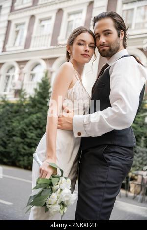 Blick aus dem niedrigen Winkel auf den bärtigen Bräutigam in Weste, der die Braut in weißem Kleid umschließt, mit Hochzeitsstrauß, Stockbild Stockfoto
