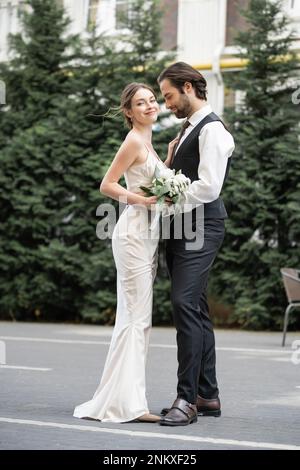Bärtige Bräutigam in Weste mit fröhlicher Braut in weißem Kleid und Hochzeitsstrauß, Stockbild Stockfoto