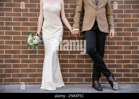 Kurzer Blick auf die junge Braut in weißem Kleid mit Hochzeitsstrauß und Hand des Bräutigams in der Nähe von Ziegelmauern, Stockbild Stockfoto