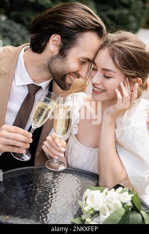 Fröhliche und junge Vermählte, die während der Hochzeitsfeier ein Glas Champagner halten, während sie lachen, Stockbild Stockfoto