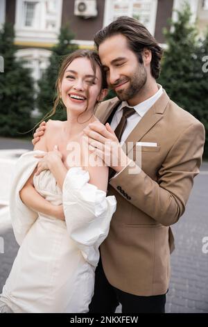 Porträt eines positiven und bärtigen Mannes, der die glückliche Braut in Hochzeitskleid umarmt, Stockbild Stockfoto