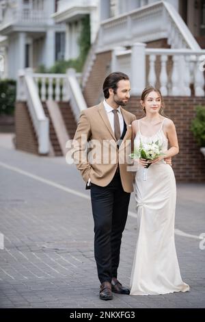 Der Bräutigam posiert mit der Hand in der Tasche, während er die Braut im Hochzeitskleid mit blühenden Blumen ansieht, Stockbild Stockfoto