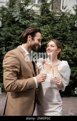 Lächelnder Bräutigam im Anzug und glückliche Braut im Hochzeitskleid, Gläser Champagner, Stockbild Stockfoto