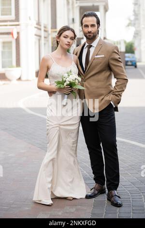 Lange Braut in Hochzeitskleid mit blühenden Blumen neben dem Bräutigam posieren mit Hand in Tasche, Stockbild Stockfoto