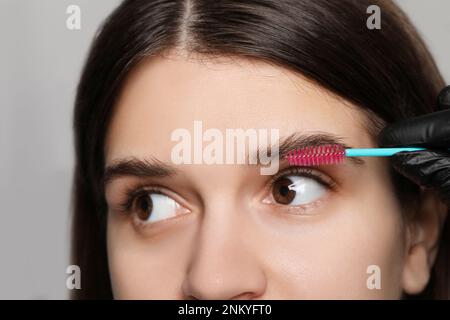 Kosmetikerin putzt die Augenbrauen von Frauen, bevor sie auf grauem Hintergrund abfärbt, Nahaufnahme Stockfoto