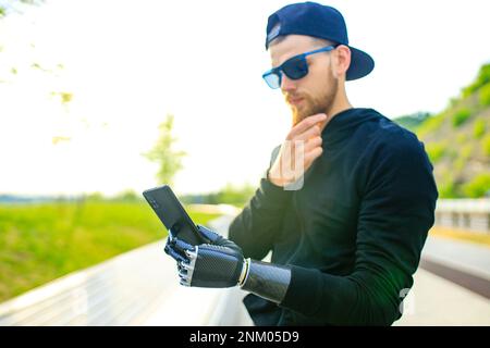 Junger bärtiger Mann mit Metallarm-Prothese im Freien Blick auf den Bildschirm des Smartphones Stockfoto