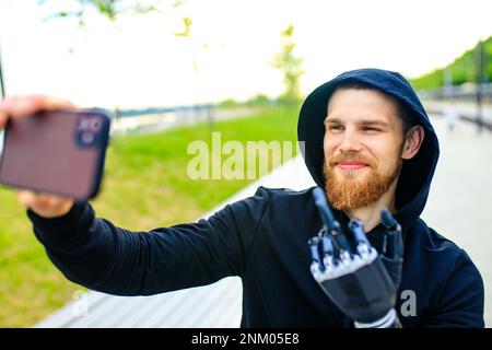 Junger bärtiger Mann mit Metallarmprothese im Freien, der Selfie-Fotos mit einer Smartphone-Kamera macht Stockfoto