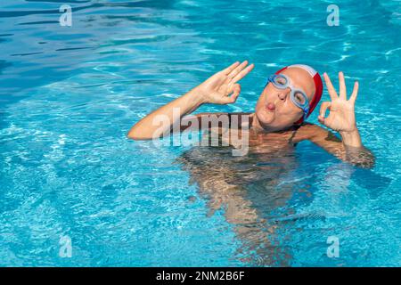 Eine weiße Frau mittleren Alters ist in einem Schwimmbecken, trägt eine Schutzbrille und eine Badekappe, steckt ihre Zunge heraus und macht das "alles in Ordnung" -Schild mit ihren Fingern Stockfoto