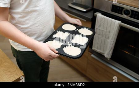 Ein Teenager mit Übergewicht hält ein Tablett mit rohen Cupcakes, Muffins, Teig. Person ohne Gesicht steht neben dem Ofen in der modernen Innenküche. Muttertag Stockfoto