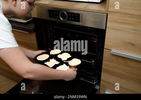 Ein übergewichtiger Teenager, der ein Tablett mit rohem Teig in den Ofen wirft, um Cupcakes und Muffins in der Küche zu backen. Muttertag, hausgemachtes Kochen, helfen mot Stockfoto