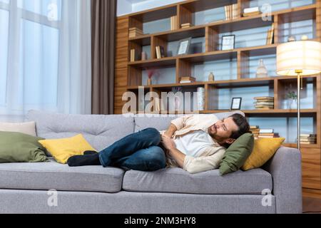 Ein junger Mann liegt zu Hause auf dem Sofa, hält seinen Magen und leidet an starken Schmerzen. Stockfoto