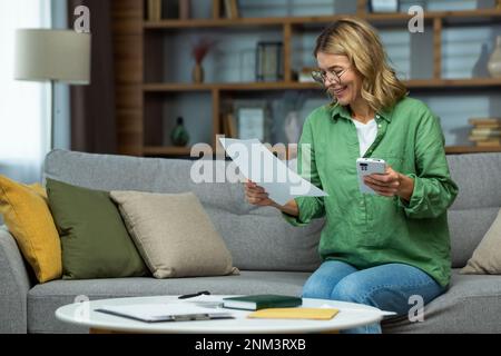 Eine ältere lächelnde oman sitzt zu Hause auf dem Sofa, hält das Telefon in der Hand, arbeitet mit Dokumenten, Finanzkonten, Miete, Darlehen, Vereinbarungen. Stockfoto