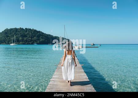 Rückansicht einer jungen asiatischen Frau in weißem Kleid, die an einem sonnigen Sommerurlaub auf einem hölzernen Pier im tropischen Meer spaziert Stockfoto