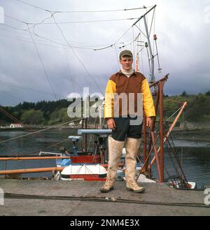 1960er, historisch, ein junger erwachsener Fischer in Wadere, Jacke und Hut, der für ein Foto auf der Slipway am Hafen von Stornoway auf der Isle of Lewis in den Äußeren Hebriden, Schottland, Großbritannien, steht Stockfoto