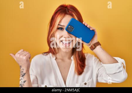 Junge weiße Frau, die das Auge mit dem Smartphone bedeckt und mit dem Daumen zur Seite zeigt, lächelt glücklich mit offenem Mund Stockfoto
