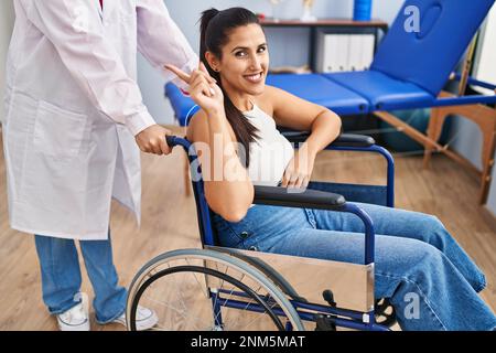 Junge hispanische Frau, die im Rollstuhl in der Physiotherapie-Klinik sitzt und glücklich lächelt, mit Hand und Finger zur Seite zeigt Stockfoto