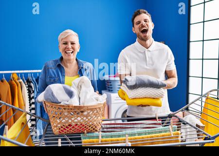 Hispanische Mutter und Sohn hängen Kleider an der Wäscheleine, lächeln und lachen laut, weil lustiger verrückter Witz. Stockfoto
