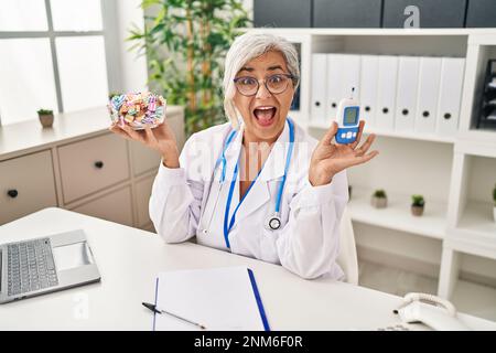 Eine Frau mittleren Alters mit grauen Haaren, die eine Arztuniform trägt und das Blutzuckermessgerät hält, die laut lächelt und lacht, weil sie einen lustigen, verrückten Witz macht. Stockfoto