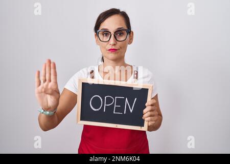Braune Frau im mittleren Alter hält ein Banner mit offenem Text und offener Hand hält ein Stoppschild mit ernsthaftem und selbstbewusstem Ausdruck, Verteidigungsgeste Stockfoto