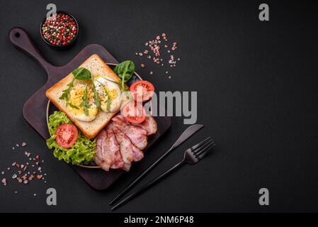 Köstliches, gesundes Mittagessen bestehend aus Speck, Toast, Eiern, Tomaten, Salat und Gemüse auf schwarzem Teller auf Betonhintergrund Stockfoto