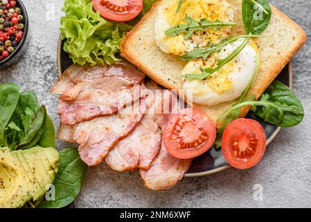 Köstliches, gesundes Mittagessen bestehend aus Speck, Toast, Eiern, Tomaten, Salat und Gemüse auf schwarzem Teller auf Betonhintergrund Stockfoto