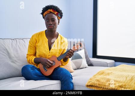 Afrikanische junge Frau, die zu Hause Ukulele spielt, skeptisch und nervös, runzelnd aufgeregt wegen des Problems. Negative Person. Stockfoto