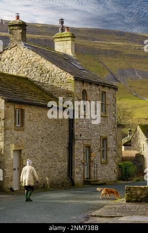 An einem sonnigen Morgen führt eine Frau ihren Hund an einem rustikalen Steinhaus mit sanften Hügeln in der Ferne vorbei, Upper Wharfedale, North Yorkshire, Großbritannien. Stockfoto