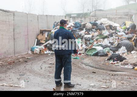 Ein Arbeiter im Recycling-Center, in dunkelblauer Arbeitskleidung, der sich einen unsortierten Müllhaufen ansieht, ist gerade in der Materialrückgewinnungsanlage angekommen, Rückansicht. Stockfoto