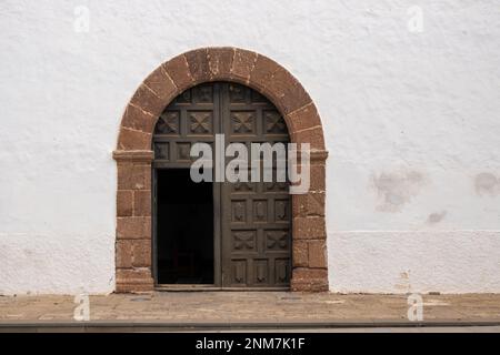 Weiße verwitterte Fassade eines Kirchengebäudes. Bogen des Seiteneingangs, mit einem Steinrahmen. Dekoratives Muster auf dem Holztor, das zum Teil offen ist Stockfoto