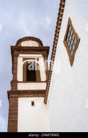 Turm einer Kirche mit Glockenturm. Weiße Fassade und braune Details. Fenster mit braunem Holzrahmen. Dachlinie. Wolkiger Himmel. Antigua, Fuerteventura, Stockfoto