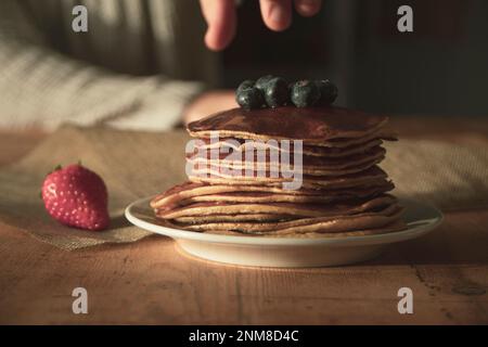 Teller mit Pfannkuchen auf einem weißen Teller mit Heidelbeeren und Erdbeeren oben in einem gut beleuchteten Setting, auf einem Holztisch und die Hand eines Mannes, der versucht zu fangen Stockfoto