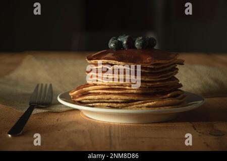 Teller mit Pfannkuchen auf weißem Teller mit Blaubeeren auf der Oberseite in gut beleuchteter Umgebung, auf Holztisch und Gabel auf der linken Seite Stockfoto