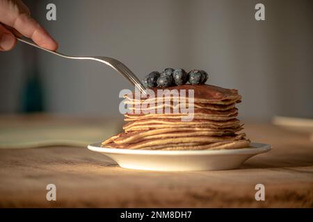 Teller mit Pfannkuchen auf weißem Teller mit Blaubeeren auf der Oberseite in gut beleuchteter Umgebung, auf Holztisch und Gabel auf der linken Seite Stockfoto