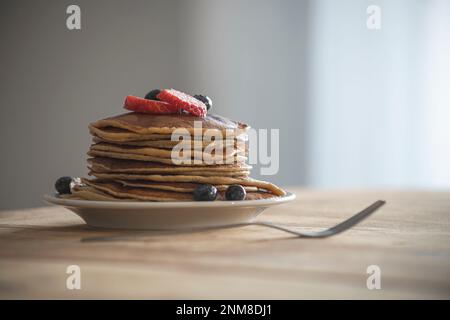 Teller mit Pfannkuchen auf weißem Teller mit Blaubeeren auf der Oberseite in gut beleuchteter Umgebung, auf Holztisch und Gabel auf der linken Seite Stockfoto