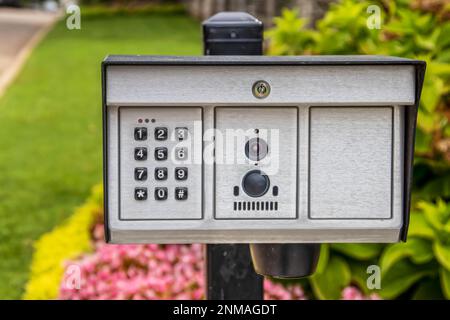 Elektrischer Toröffner mit Tastenfeld und Kamera und Mikrofon im Außenbereich des Anwesens mit wunderschönen verschwommenen Blumen im Hintergrund Stockfoto