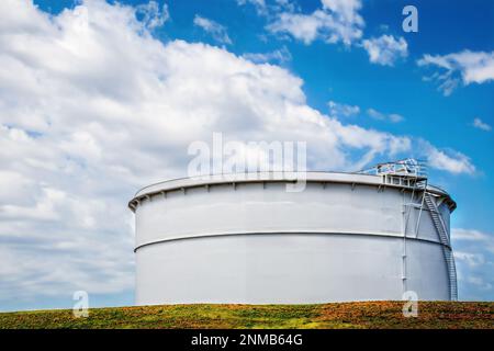 Großer, weißer Petrochemie-Lagertank am Terminal vor blauem, wolkigen Himmel mit farbenfrohem Gras und rotem Ton im Vordergrund Stockfoto