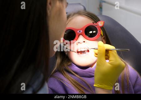 Zahnfüllungen bei Kindern Milchzähne verschlechtern sich schnell Zahnarzt bläst Luft auf Kariesloch und schaut durch mit einem Spiegel Mädchen sitzt in einem Zahnstuhl in Sonnenbrille junge, wunderschöne echte Menschen Stockfoto