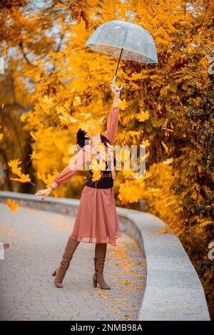 Hübsches Mädchen in einem Kleid mit Schirm im Herbstpark. Sie hält ihn über den Kopf, die Herbstblätter fallen aus ihm heraus Stockfoto