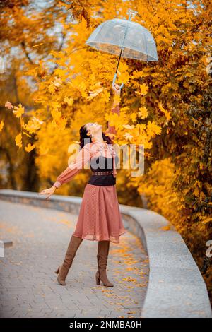 Hübsches Mädchen in einem Kleid mit Schirm im Herbstpark. Sie hält ihn über den Kopf, die Herbstblätter fallen aus ihm heraus Stockfoto