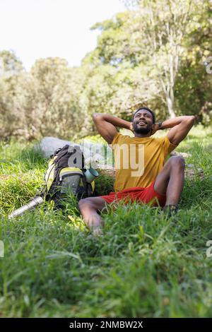 Lächelnder afroamerikanischer Mann, entspannt, sitzt im sonnigen Wald mit Rucksack, Kopierraum Stockfoto