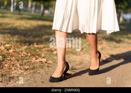 Eine Frau mit weißem Faltenrock und schwarzen Wildlederstilettos, die nach der Arbeit im Herbstpark spaziert. Stockfoto