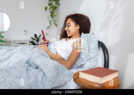 Glückliche junge lateinamerikanische Frau, die morgens ihr Handy im Bett benutzt Stockfoto