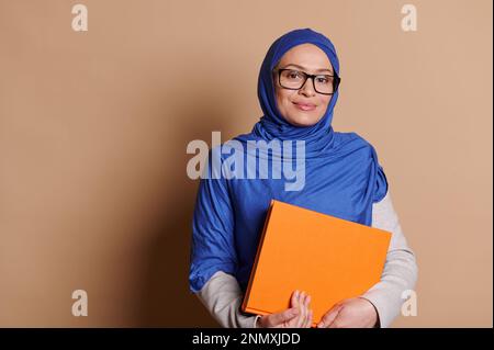 Selbstbewusste, reife muslimische Lehrerin in einem Hidschab, schaute in die Kamera, posierte mit einem Stapel Bücher, auf beigem Hintergrund Stockfoto