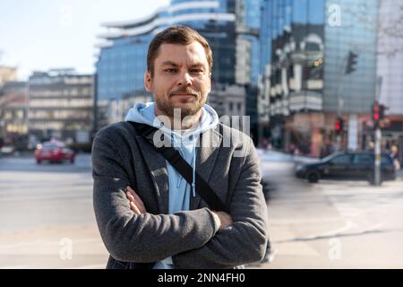 Ein Mann mit gekreuzten Armen, der auf der Straße gegen Gebäude der Stadt steht und in die Kamera schaut. Stockfoto